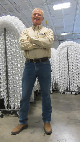 A man standing confidently with arms crossed in a warehouse setting, surrounded by large stacked objects, possibly mattresses or components, symbolizing the production process behind a tractor trailer mattress.